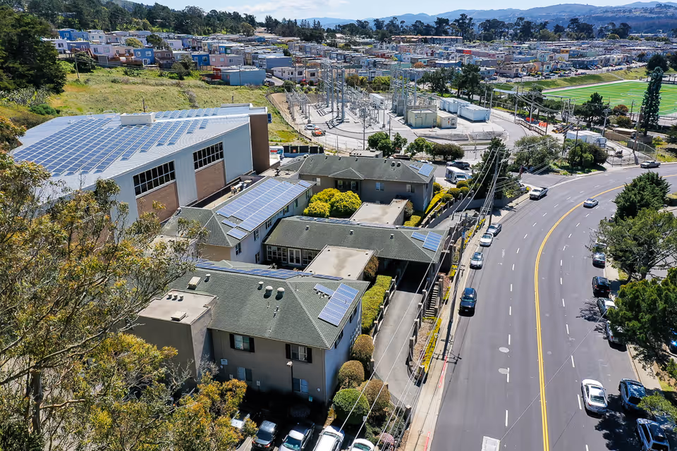 Aerial view of a senior living facility with multiple buildings featuring solar panels on the roofs, adjacent to a curved road with parked cars. In the background, there is a substation, residential houses, and a sports field surrounded by trees and hills.