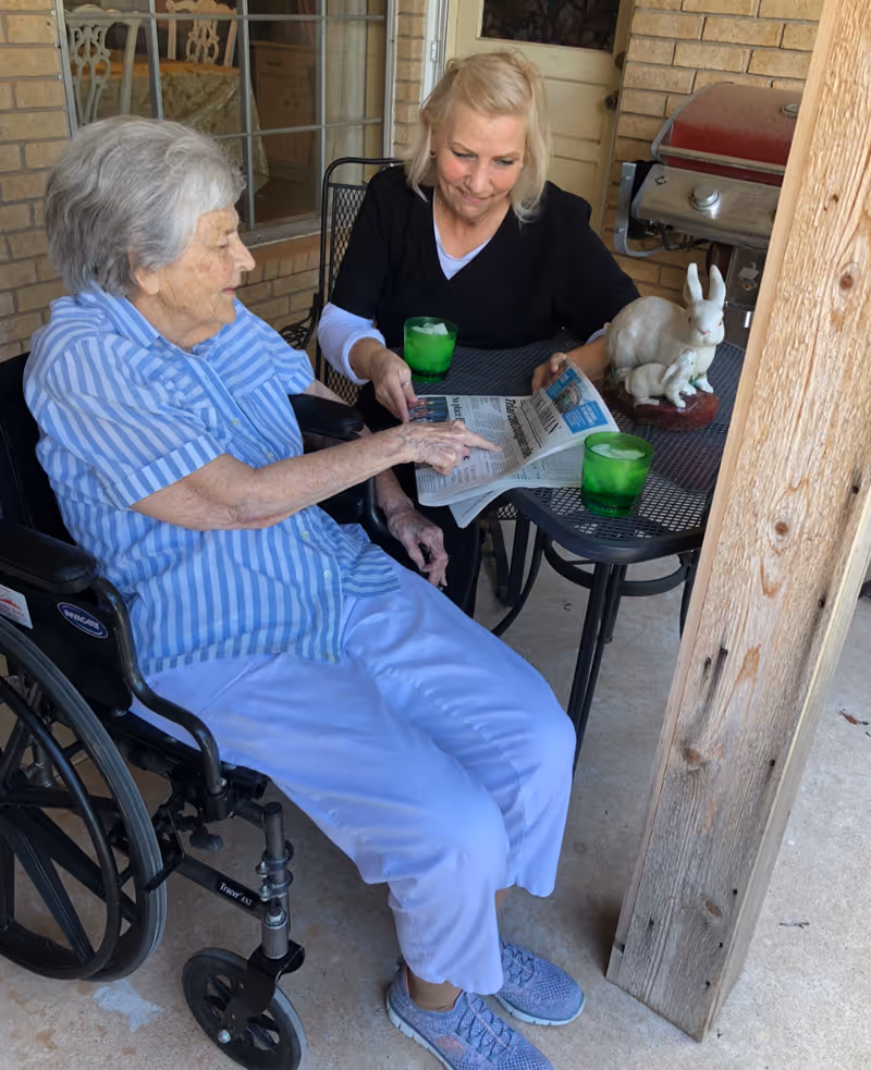 An elderly woman in a wheelchair wearing a blue striped shirt and light blue pants is sitting next to a woman in black scrubs. They are sitting at a small outdoor table with two green glasses and a white rabbit figurine. The elderly woman is pointing at a newspaper that the woman in scrubs is holding. They appear to be on a covered patio with a brick wall and a barbecue grill in the background.