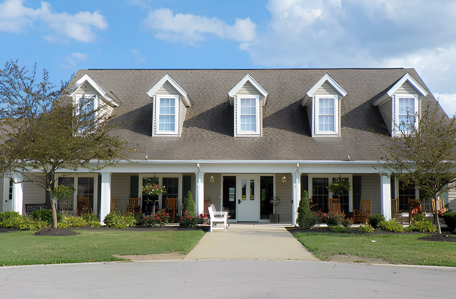 Front exterior of a two-story residential-style building with a covered porch, dormer windows, rocking chairs, and landscaped lawn.