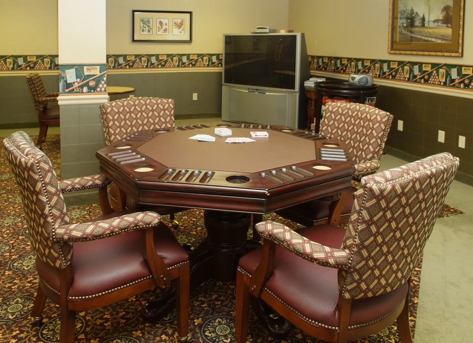 Carpeted senior community interior with an octagonal card table surrounded by upholstered chairs and a television in the background.