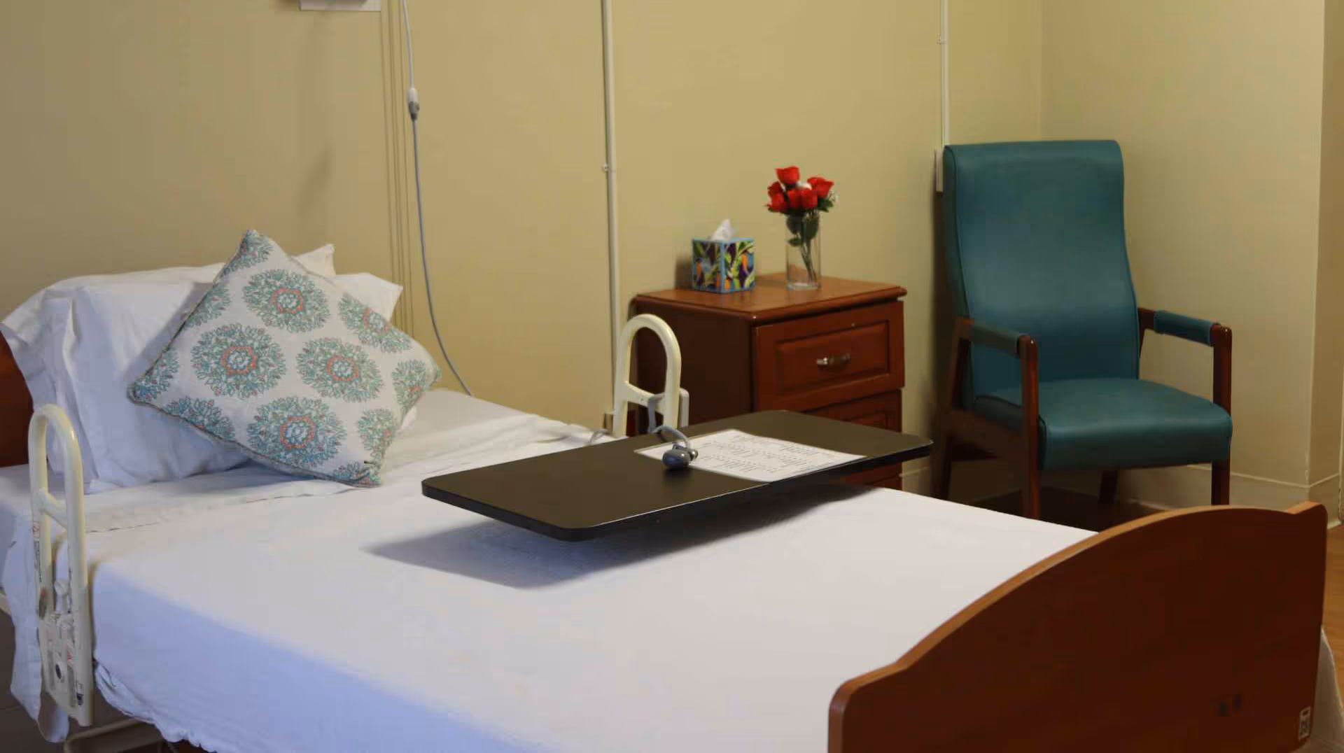 A neatly made patient bed with a patterned pillow and overbed table, a nightstand with flowers, and a teal armchair in a room.