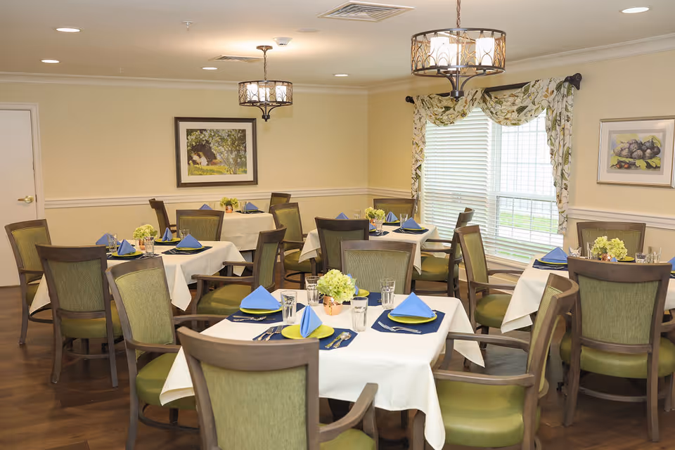 A dining room with several tables covered in white tablecloths, each set with blue napkins, plates, glasses, and silverware. The room has green cushioned chairs, two hanging light fixtures, framed artwork on the walls, and a large window with floral curtains letting in natural light.