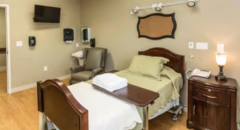 A senior facility bedroom with a wooden bed, nightstand, chair, and mounted TV.