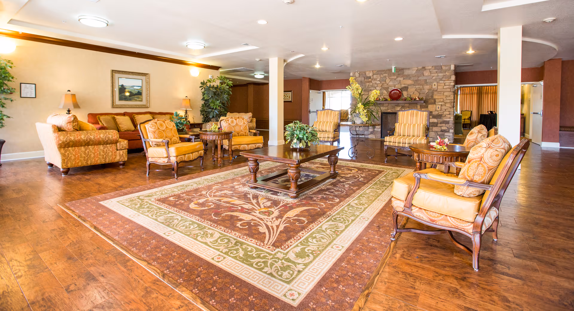 Spacious furnished common living room with armchairs, sofas, coffee tables, a patterned area rug, and a stone fireplace.