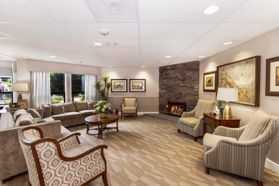 A cozy living room in a retirement and assisted living facility featuring a stone fireplace with a fire burning, multiple upholstered armchairs and sofas arranged around a wooden coffee table with a floral centerpiece, large windows with white curtains letting in natural light, framed artwork on the walls, and a table lamp on a side table.