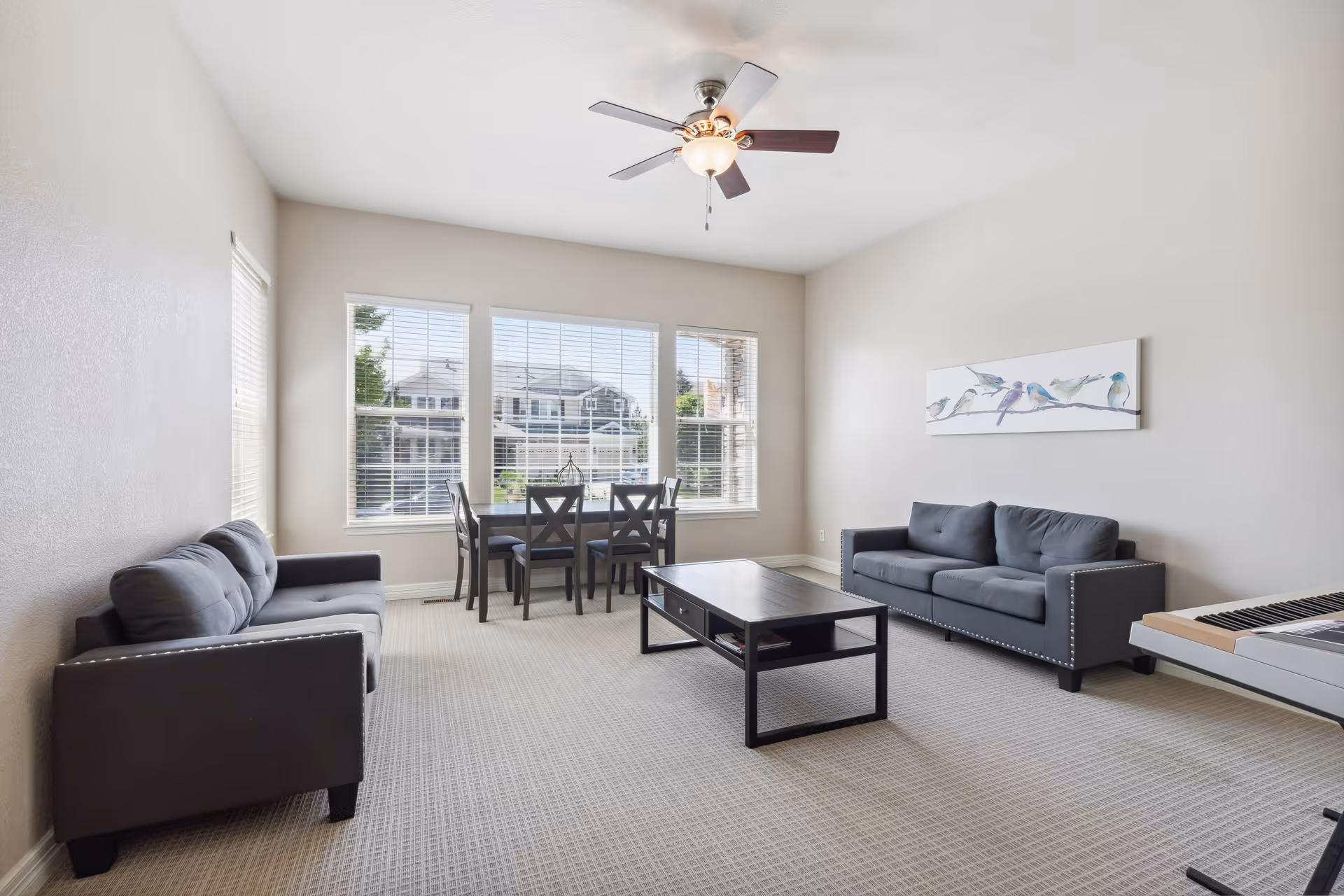 Bright living room with two gray sofas, a black coffee table, a dining table by large windows, and a ceiling fan.
