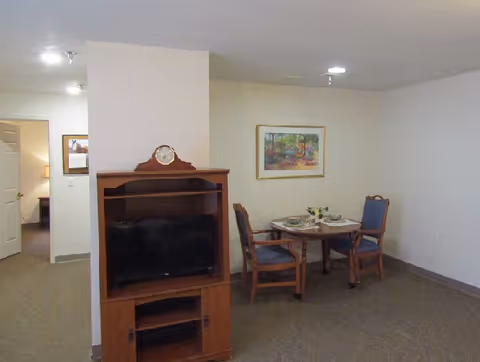 Interior view of a room in a senior living facility showing a wooden TV stand with a clock on top and a flat-screen TV. To the right, there is a small round dining table set with two chairs and place settings. A framed painting hangs on the wall above the table. The room has beige walls and carpeted floor.