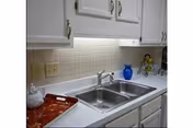 A clean kitchen countertop with a double stainless steel sink, white cabinets above and below, a beige tiled backsplash, a red tray on the left side, a blue pitcher, and a decorative fruit holder on the right side.