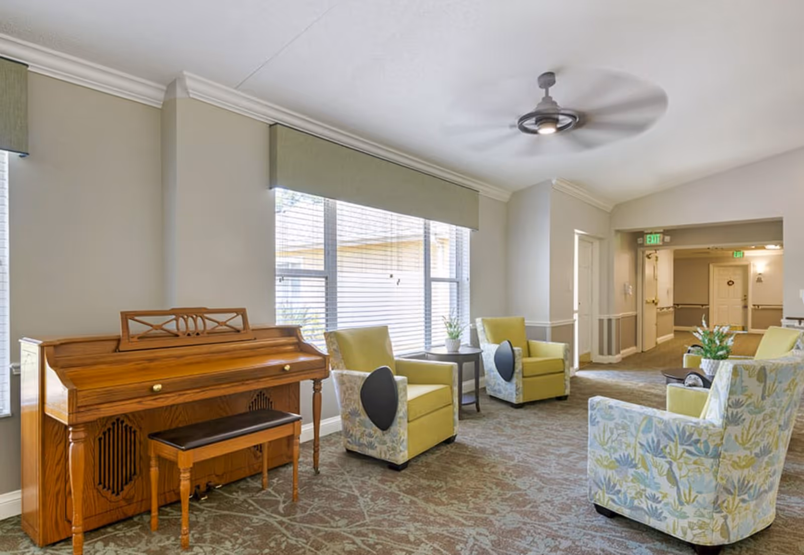 Bright communal lounge with a wooden piano, several upholstered armchairs and side tables arranged by large windows.