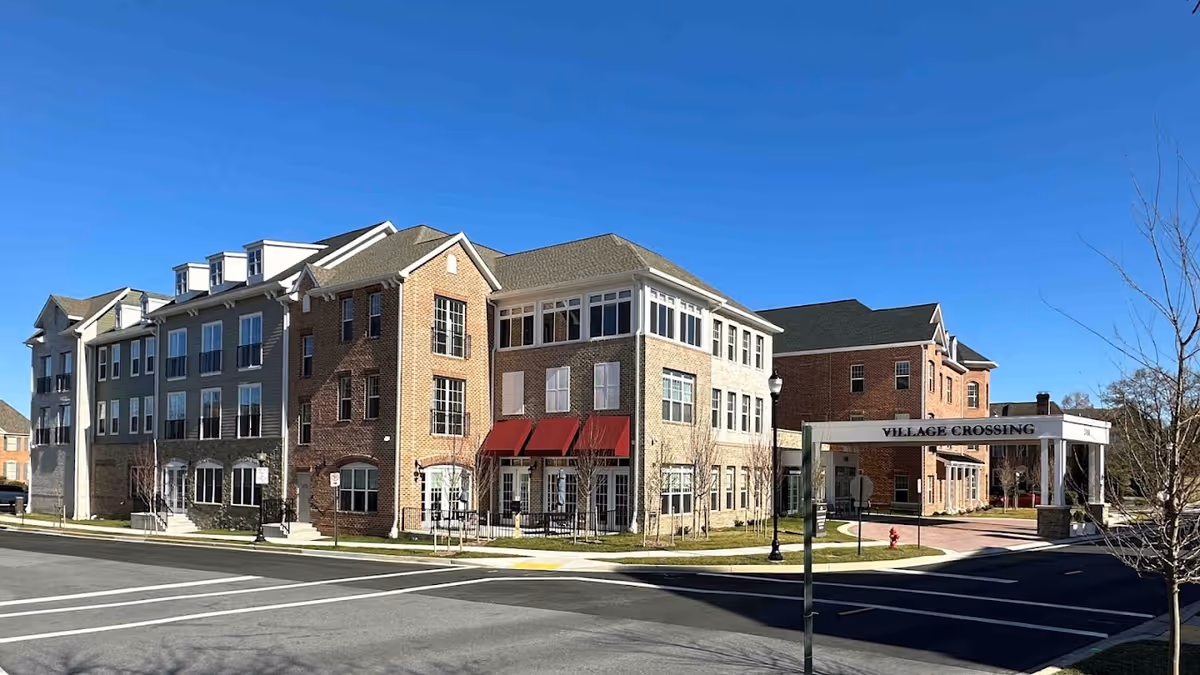Exterior view of a multi-story senior living facility building with brick and stone facades under a clear blue sky. The entrance canopy displays the name 'Village Crossing'. The surrounding area includes a paved road, sidewalks, and some leafless trees.