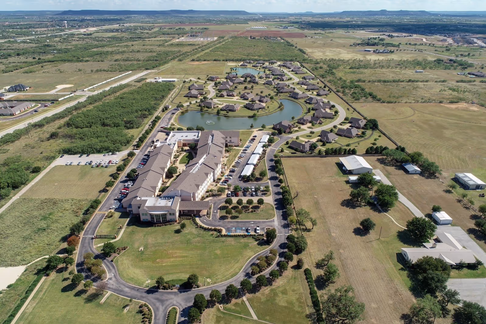 Aerial view of Wesley Court senior living facility showing a large main building with parking lots, surrounded by smaller residential houses arranged around a pond with fountains. The facility is set in a rural area with open fields and scattered trees.