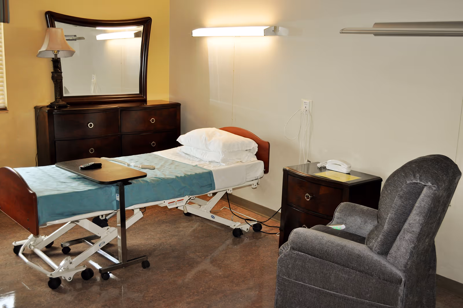 A room with a hospital-style adjustable bed covered with a teal blanket and white pillows, a dark wooden dresser with a mirror and a lamp on top, a dark wooden nightstand with a white telephone, and a gray upholstered recliner chair.