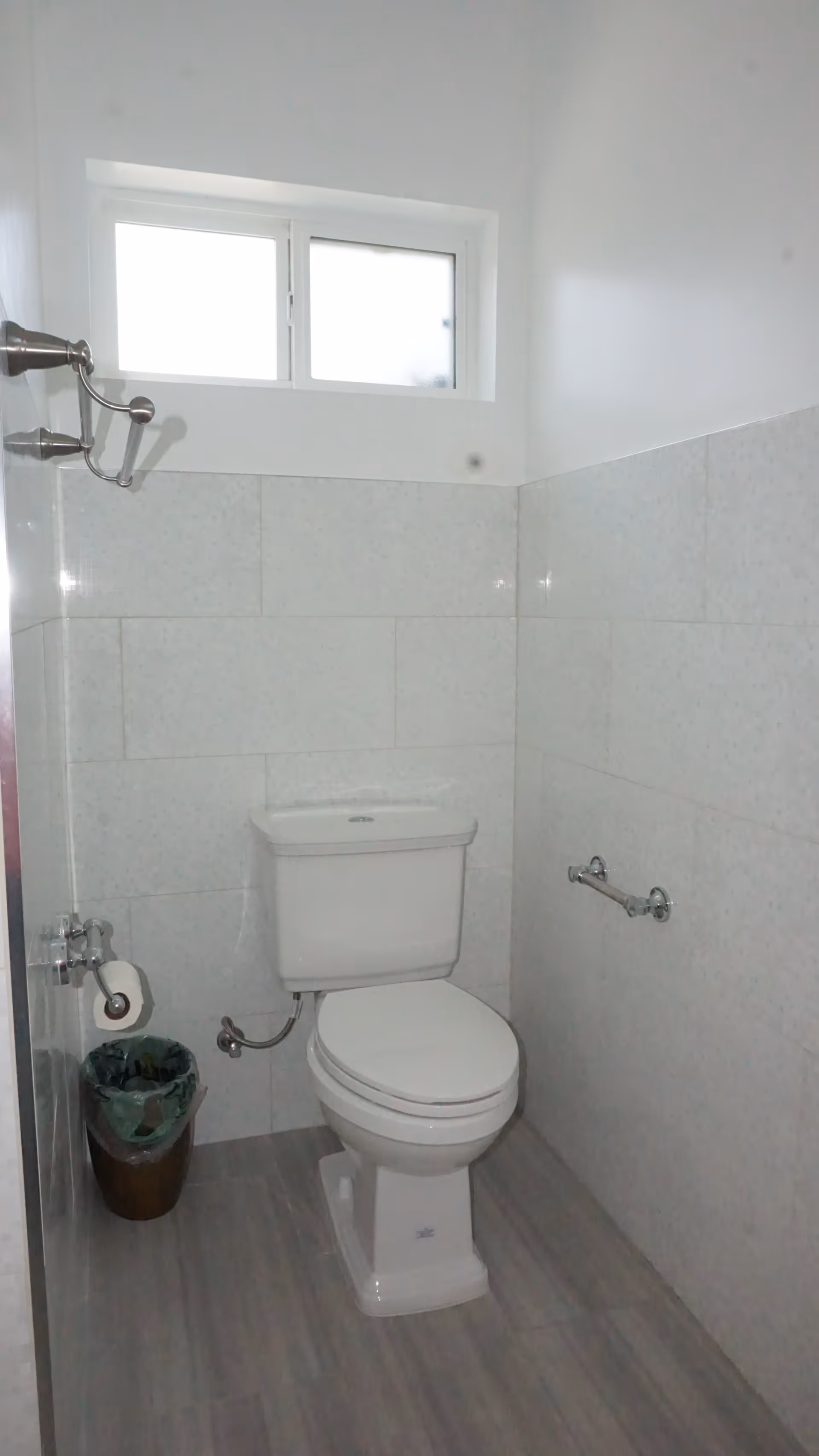 A small bathroom with a white toilet positioned against a tiled wall. There is a window above the toilet letting in natural light. A toilet paper holder with a roll of toilet paper is mounted on the left wall, and a small trash bin is placed on the floor next to it. The floor has light-colored tiles, and there is a towel hook on the left wall.