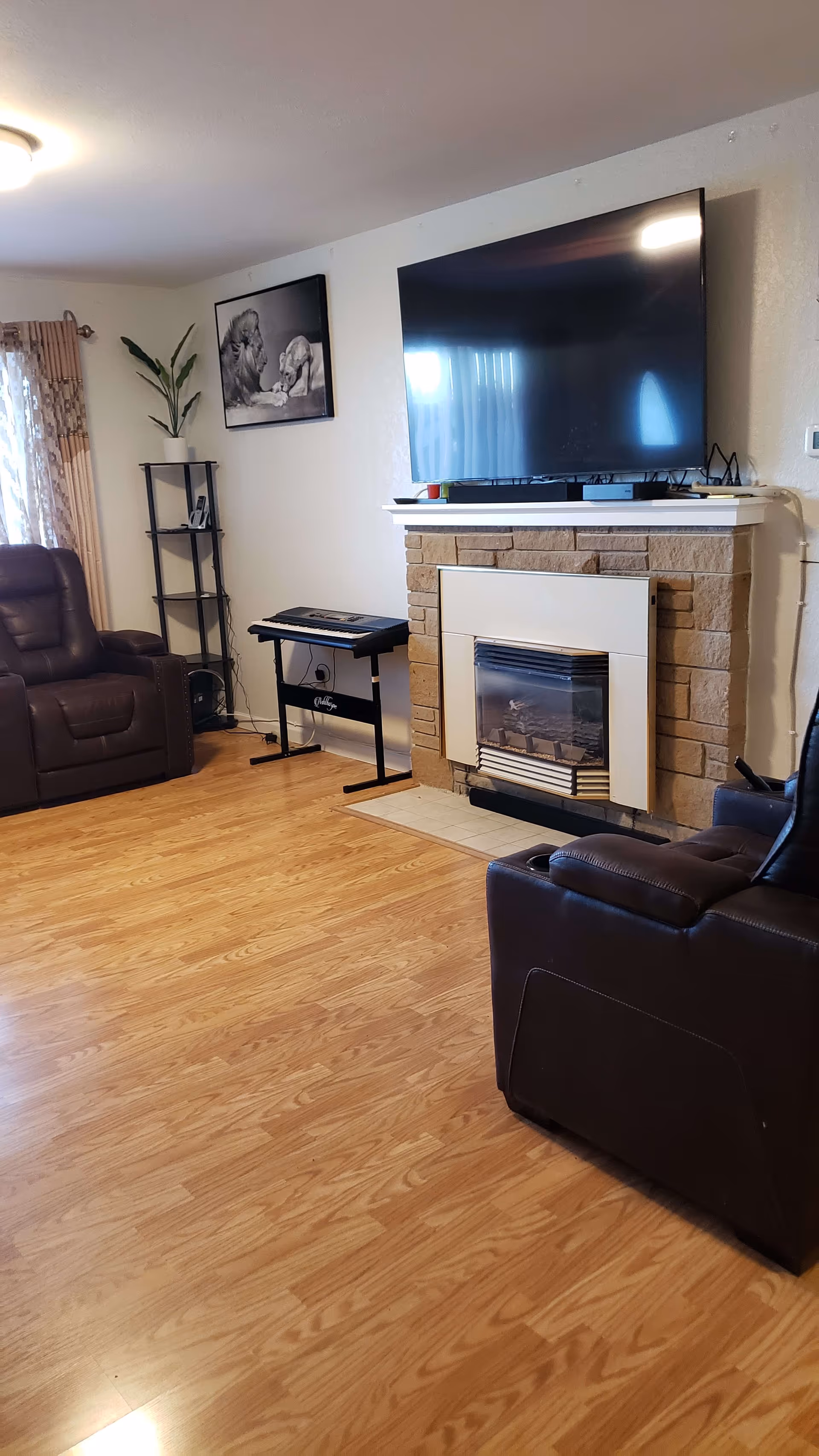 A living room with wooden flooring, two brown leather recliners, a fireplace with a mounted flat-screen TV above it, a black electronic keyboard on a stand, a black shelving unit with a potted plant and a phone, and a framed black and white picture of a lion and a dog on the wall.
