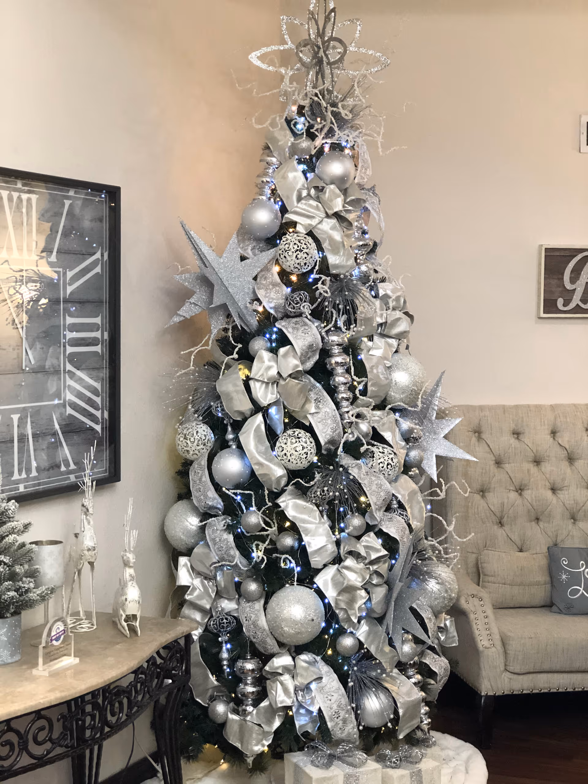 A decorated Christmas tree with silver and white ornaments, ribbons, and lights stands next to a beige tufted armchair in a cozy living room. A large wall clock and small decorative items are on a table nearby.