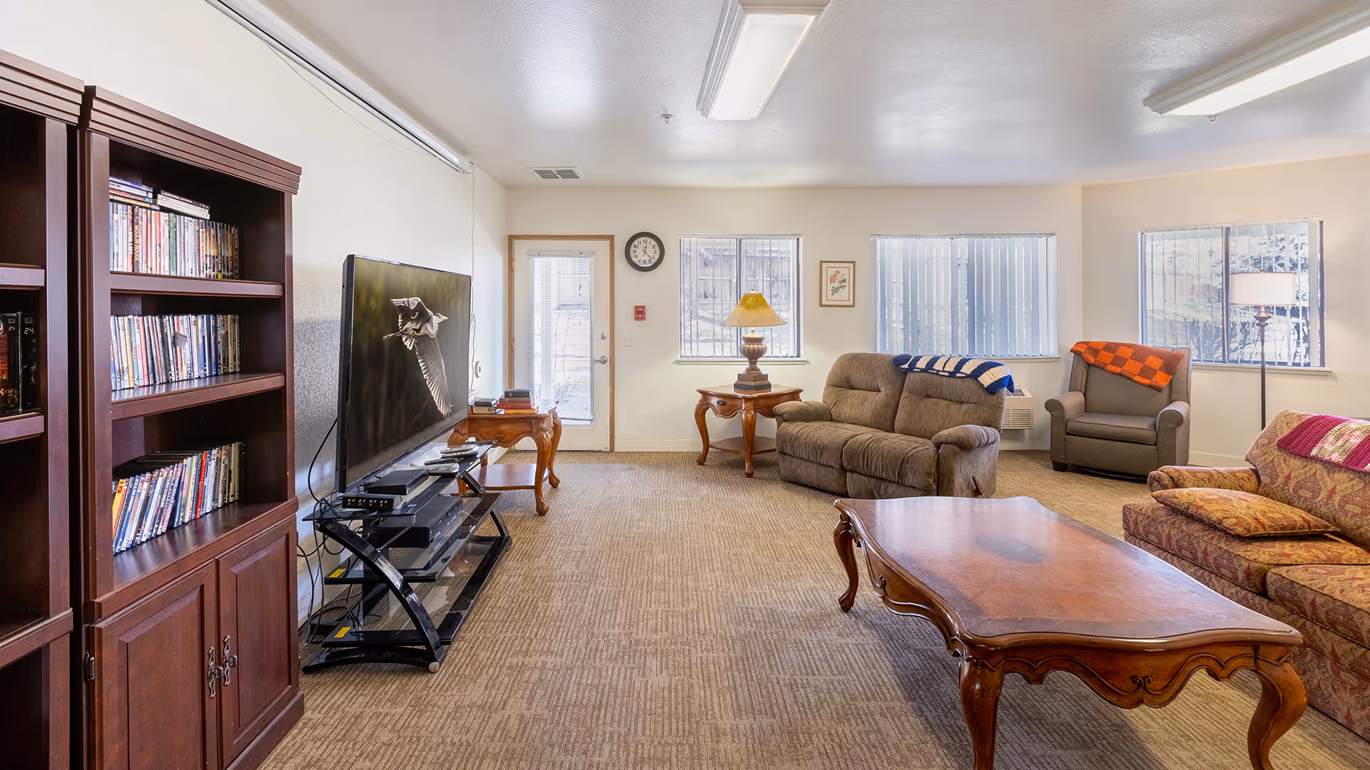 A cozy living room in a senior living facility featuring a large flat-screen TV on a black stand, a wooden bookshelf filled with DVDs, a brown loveseat and armchair with colorful blankets, a patterned sofa, a wooden coffee table, side tables with lamps, and windows with vertical blinds letting in natural light.