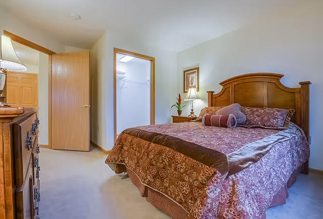 A bedroom with a wooden bed frame and a bed covered in a patterned maroon and brown bedspread with matching pillows. There is a wooden nightstand with a lamp and a framed picture on the wall above it. The room has beige carpet and light-colored walls. An open door reveals a closet with white walls and a wire shelf. Another door is partially open, showing a wooden door and part of another room.