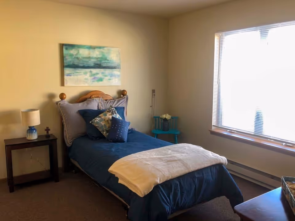 A cozy bedroom with a single bed made with blue bedding and multiple pillows. There is a small wooden nightstand with a lamp and a small cross on it to the left of the bed. A blue chair with a vase of flowers is placed near a window with blinds letting in natural light. A painting with blue and green hues hangs on the wall above the bed.
