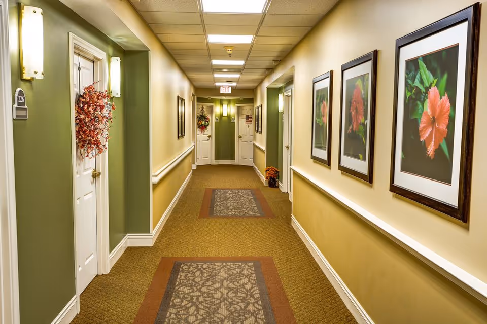 Carpeted interior hallway in a senior living facility with doors, framed floral pictures, and decorative wreaths.