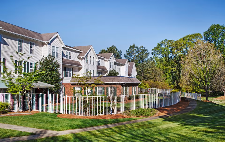 Exterior view of a multi-story senior living facility with white siding and brick accents, surrounded by a white metal fence. There is a well-maintained lawn, trees, and a paved walkway curving around the building under a clear blue sky.