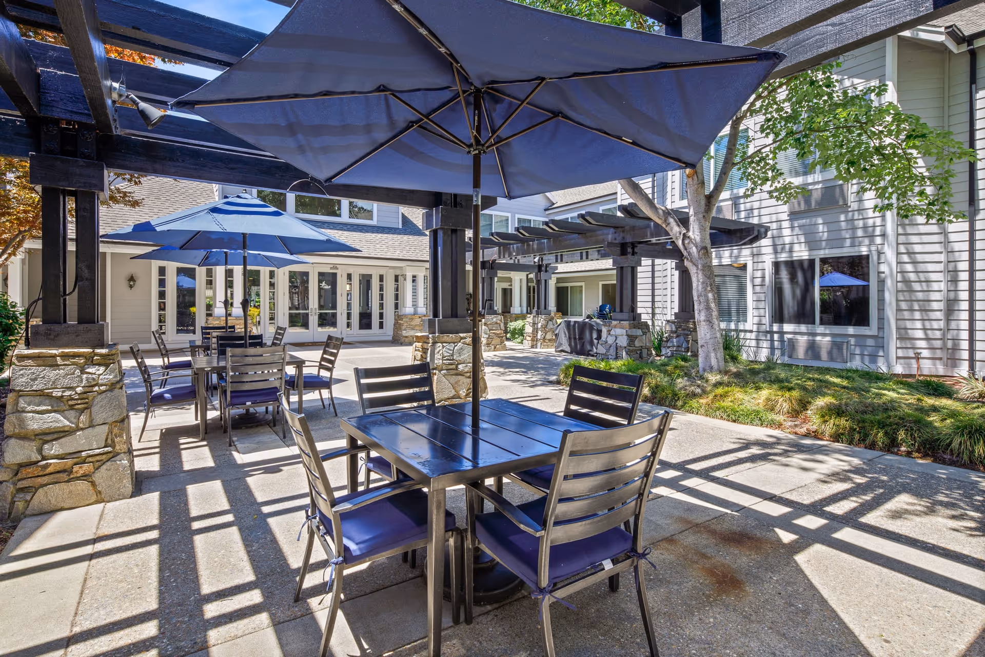 Outdoor patio area with multiple tables and chairs under large blue umbrellas. The patio is surrounded by a building with large windows and stone pillars supporting wooden pergolas. There are trees and greenery around the patio, and the area is well-lit with natural sunlight.