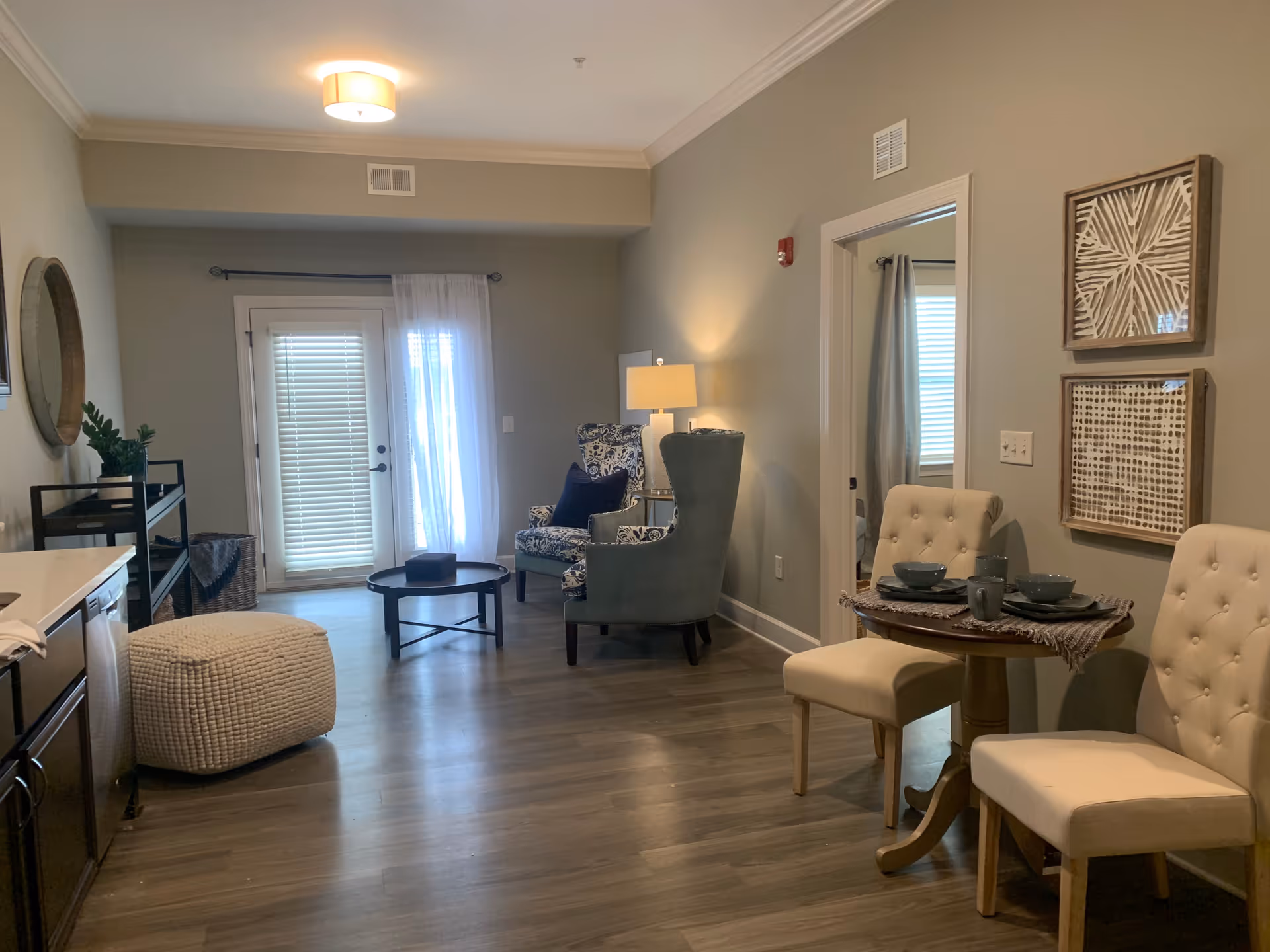 A cozy living area with light gray walls and wooden flooring. The room features a small round dining table with two beige tufted chairs set with bowls and mugs. To the right, there are two framed abstract artworks on the wall. In the background, there are two armchairs, one patterned and one solid gray, with a floor lamp between them. A round coffee table and a white knitted pouf are also visible. French doors with blinds and sheer curtains let in natural light.