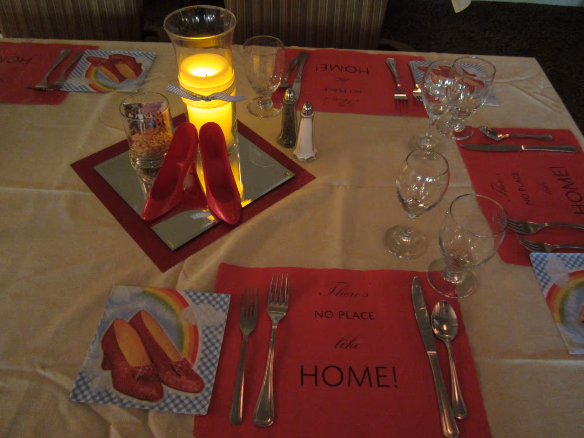 A dining table set with red placemats that say 'There's NO PLACE like HOME!', silverware, glasses, and napkins featuring red shoes and a rainbow. The centerpiece includes a lit candle in a glass holder, a small glass with popcorn, and a pair of small red shoes on a mirrored square.