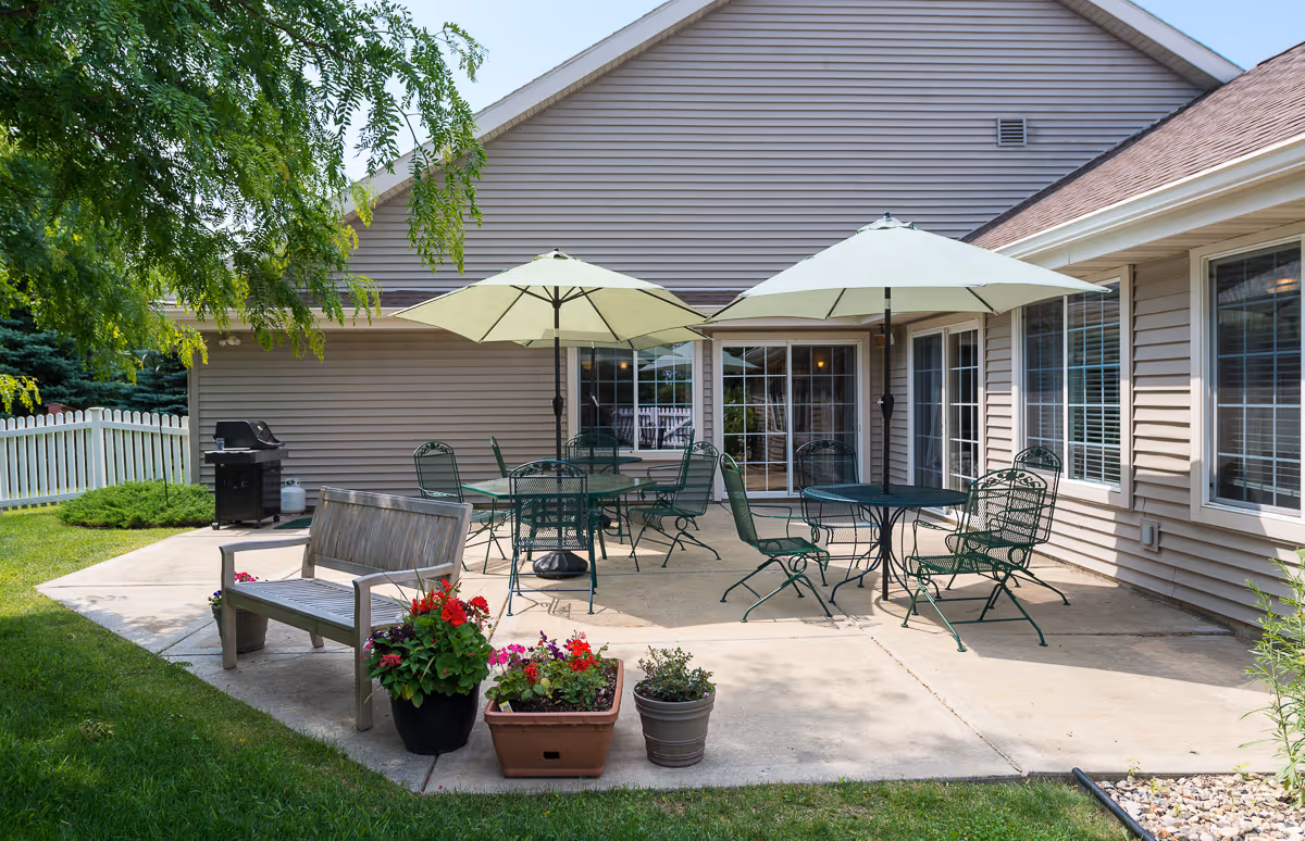 Outdoor patio area with metal tables and chairs under large umbrellas, a wooden bench, potted flowers, a barbecue grill, and a beige building with multiple windows and sliding glass doors.