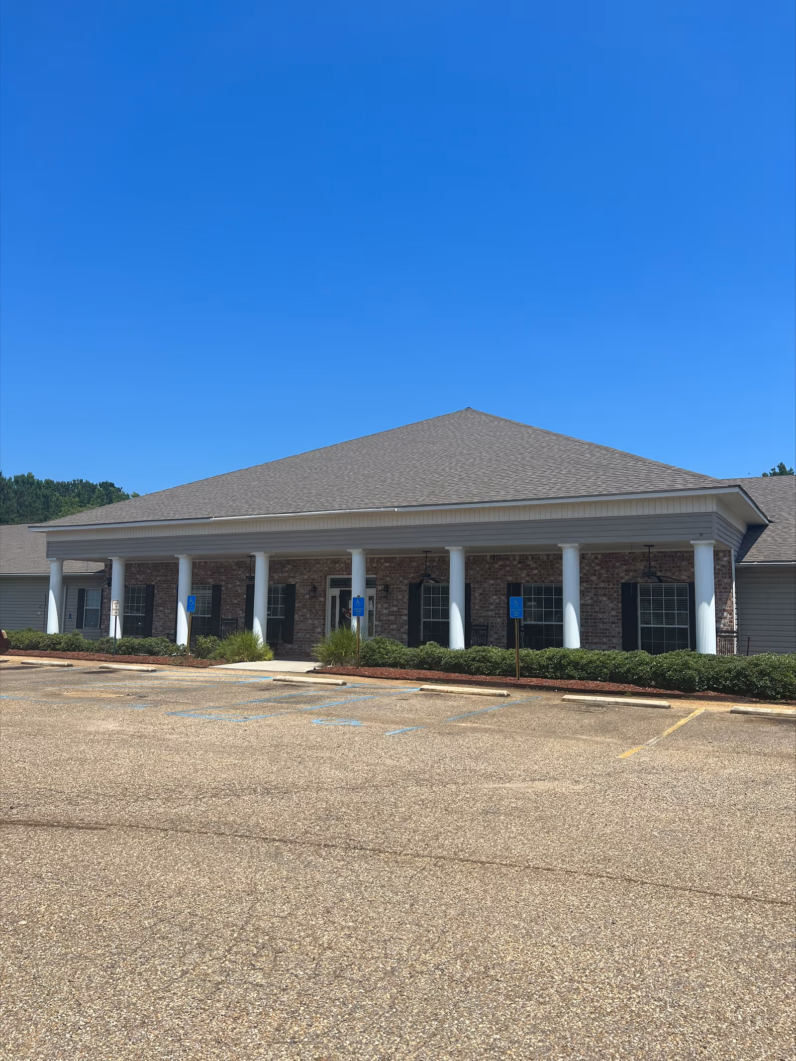 Exterior view of a single-story brick building with white columns and a gray shingled roof under a clear blue sky. The building has several windows with black shutters and a covered entrance. There is a parking lot with marked handicap spaces in front of the building.