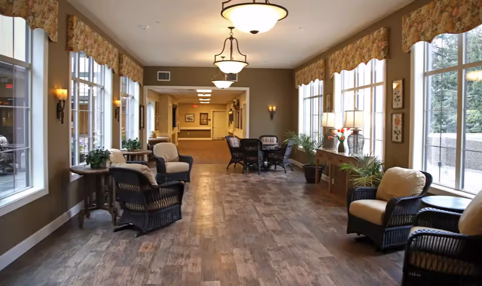 A bright, spacious interior hallway with large windows on the right side letting in natural light. The hallway is furnished with wicker chairs with cushions, small tables with plants, and a console table with lamps and flowers. The floor is wood-patterned, and the walls are painted in warm tones with decorative wall sconces and framed artwork.