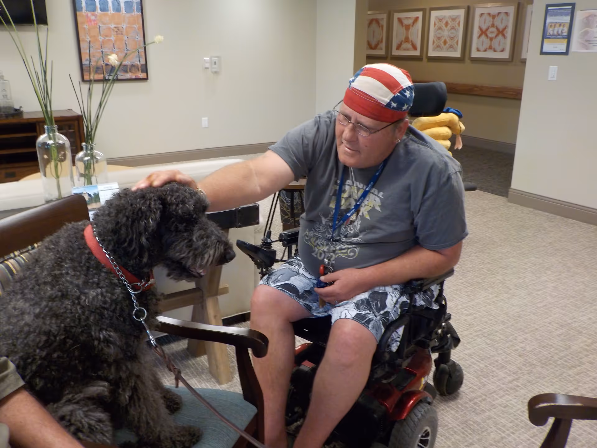 A man in a wheelchair wearing a red, white, and blue bandana and casual clothes is petting a large black curly-haired dog sitting on a chair in a room with beige walls and carpet. There are framed artworks on the wall and decorative vases with tall plants on a table in the background.