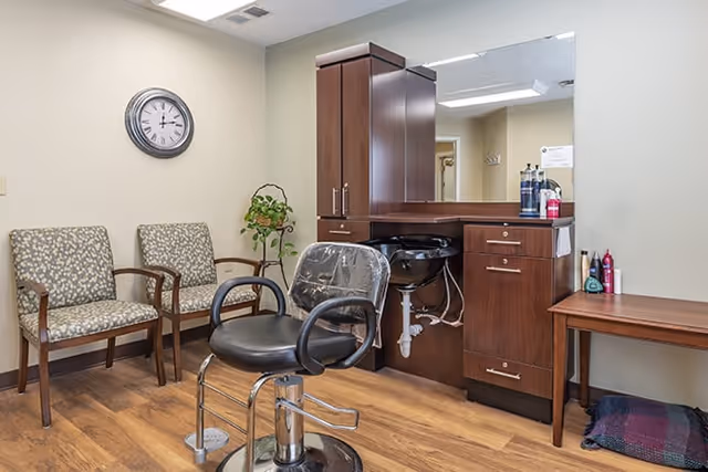 Interior of a salon area in a senior living facility with a black salon chair in front of a wooden cabinet and sink. Two patterned armchairs are placed against the wall under a round clock. A small plant is on a stand between the chairs and the cabinet. The floor is wooden and the walls are light-colored.