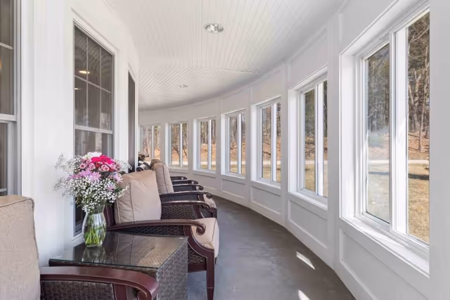 A bright, curved indoor sunroom with multiple large windows letting in natural light. The room features cushioned wicker chairs lined up along the wall, with a glass-top side table holding a vase of pink and white flowers. Outside the windows, a wooded area is visible.