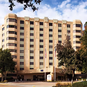 Exterior view of a multi-story senior living facility building with beige and brown vertical stripes, several windows, and an entrance canopy. There are trees and a driveway in front of the building under a partly cloudy sky.