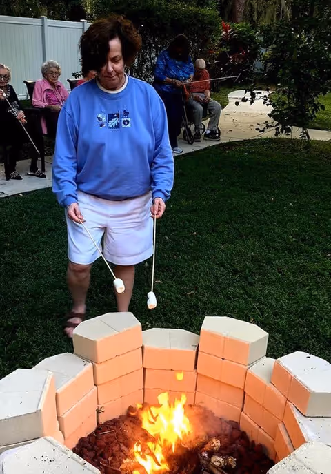 A woman in a blue sweatshirt and white shorts roasting marshmallows over a fire pit made of light-colored bricks in a grassy outdoor area. In the background, several elderly people are seated along a white fence, some using walkers or wheelchairs.