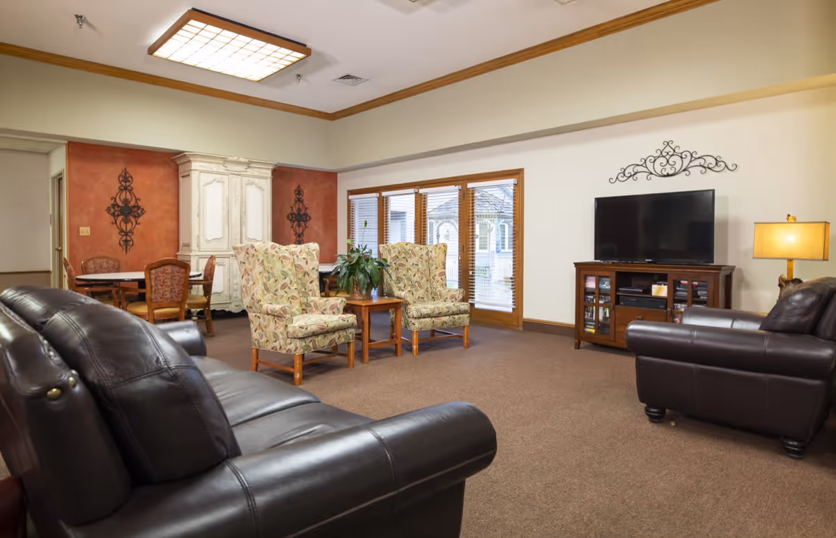 A cozy living room in an assisted living facility featuring two dark leather sofas, two floral upholstered armchairs, a wooden coffee table with a plant, a wooden TV stand with a flat-screen TV, a floor lamp, and a dining area with a table and chairs in the background. The room has beige walls, carpeted floor, and large windows with wooden blinds.