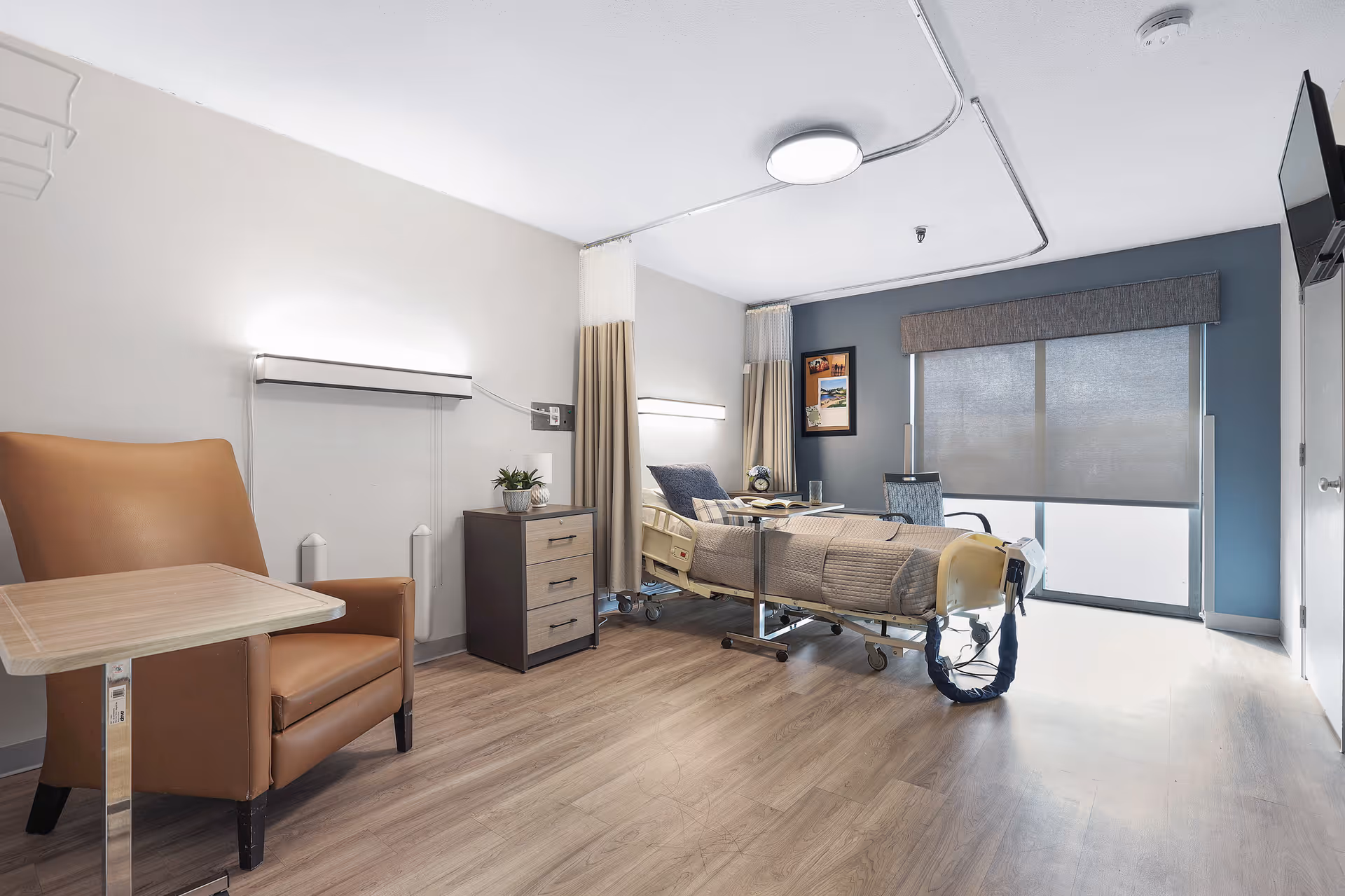A clean and modern healthcare facility room with a hospital bed, a brown leather armchair with a small table, a bedside cabinet with a plant, a window with blinds, and a wall-mounted TV.