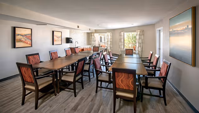 A bright dining room with multiple rectangular tables and upholstered chairs, wall art, and windows letting in natural light.