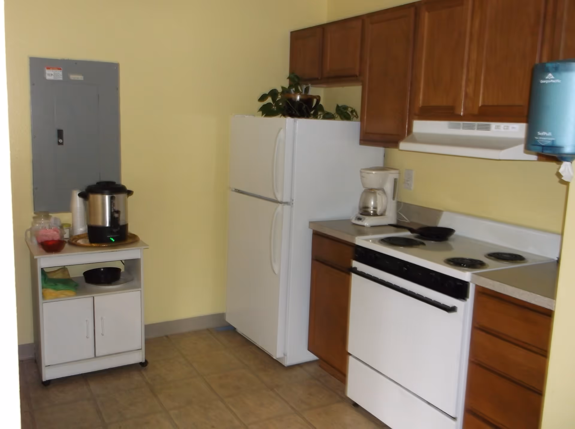 A small kitchen area with a white refrigerator, white electric stove with four burners, wooden cabinets, a coffee maker on the counter, and a small white cart holding a coffee urn, cups, and other items. The walls are painted yellow and the floor is tiled.