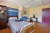 Single-occupancy senior living bedroom with a hospital-style bed, blue chair, side table, dresser, and wood flooring.