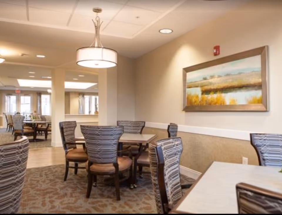 Dining area with round tables and upholstered chairs under pendant lighting and a landscape painting on the wall.