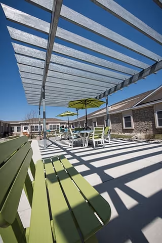 Sunlit outdoor courtyard with green benches, tables and umbrellas under a pergola between single-story building wings.