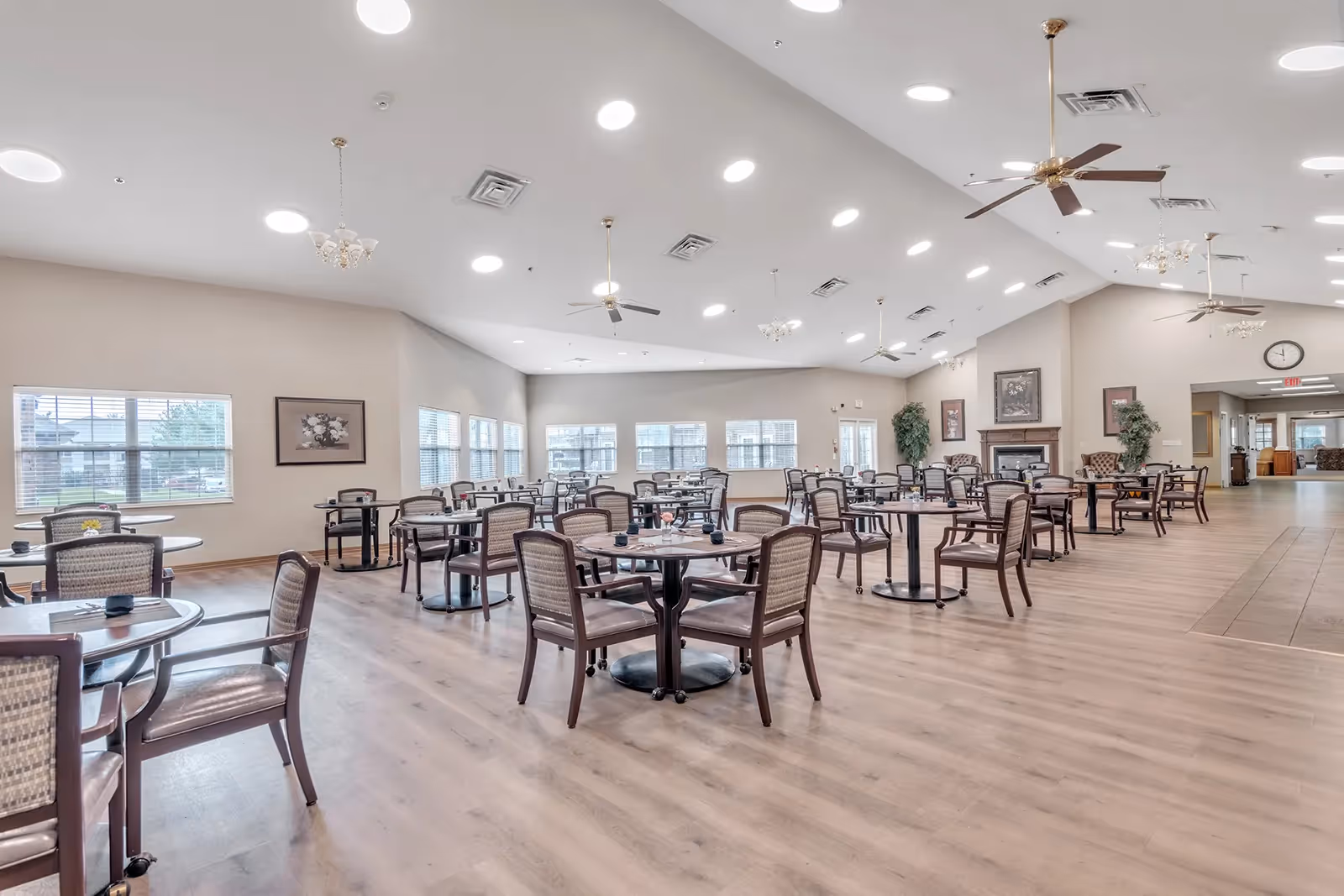A spacious dining room with multiple round tables and wooden chairs arranged neatly on a light wood floor. The room has large windows letting in natural light, ceiling fans, and recessed lighting. There is a fireplace with framed artwork above it and potted plants near the walls.