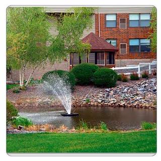 A landscaped outdoor area with a small pond featuring a water fountain, surrounded by green grass, bushes, trees, and a rocky edge. In the background, there is a brick building with windows and a small gazebo-like structure.