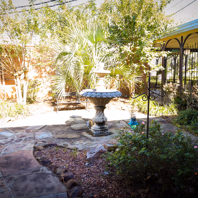 A sunny outdoor garden area with a stone pathway leading to a decorative stone fountain surrounded by plants and trees. There is a metal bench in the background and a covered patio area with black metal railings on the right side.