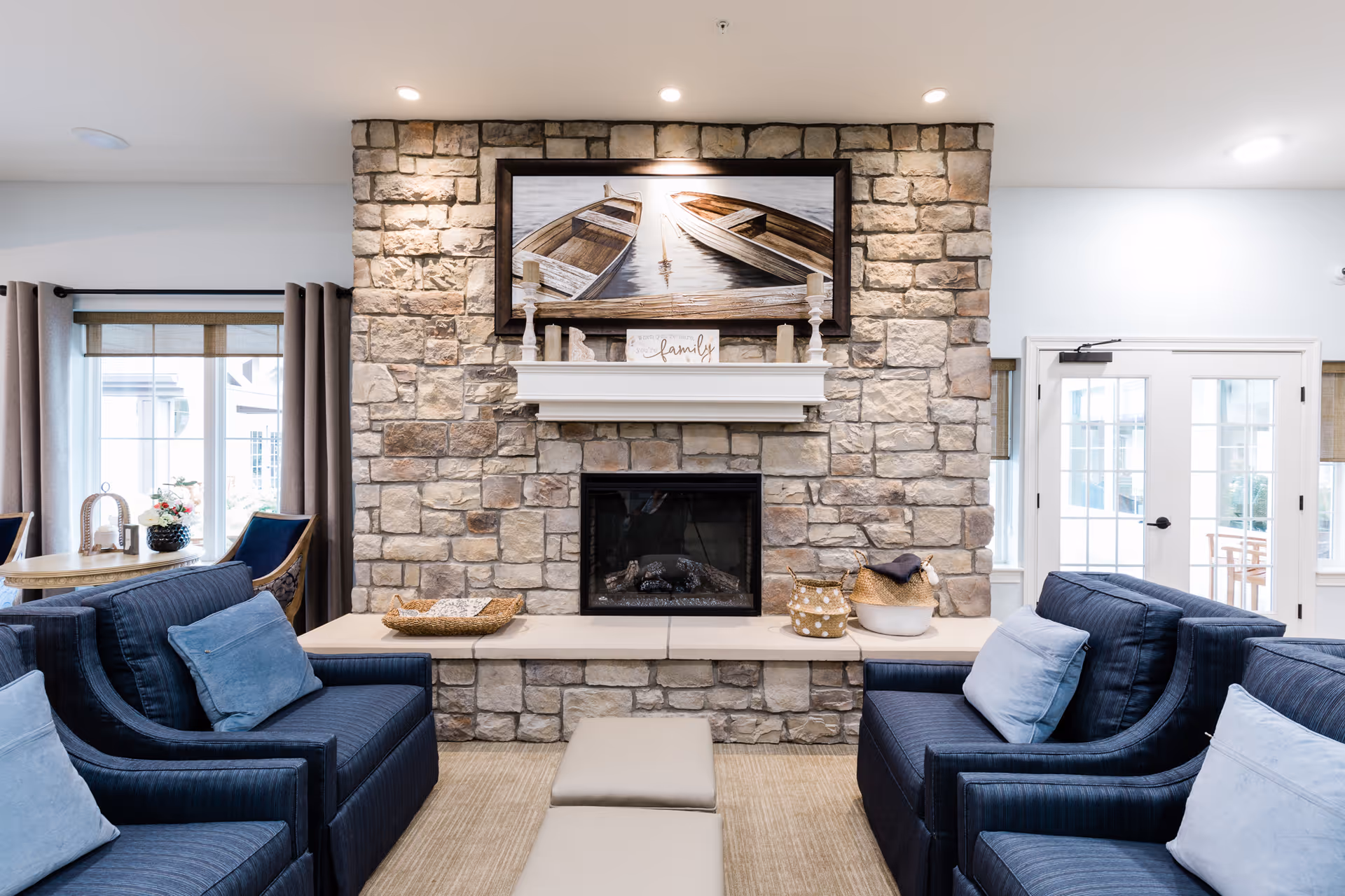 Cozy seating area with navy armchairs facing a stone fireplace topped with a mantel and framed boat artwork.