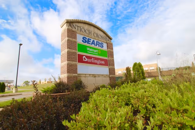 Large outdoor shopping center sign for Antioch Crossing displaying store names Sears, Walmart Neighborhood Market, and Burlington, surrounded by greenery and under a partly cloudy sky.