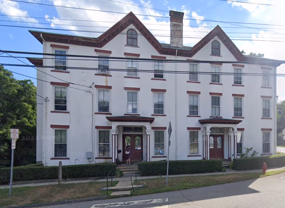 Front exterior view of a three-story white building with red trim around the windows and doors. The building has two main entrances with small covered porches and a large chimney in the center. There are bushes along the front and a sidewalk leading to the entrances. A person is standing near one of the doors. Power lines run across the top of the image.