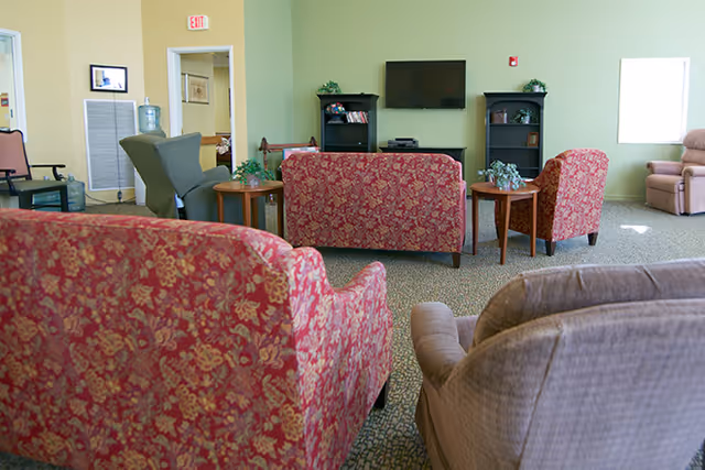 A common area in The Chesterley featuring multiple upholstered chairs and sofas arranged around small wooden tables. A flat-screen TV is mounted on a light green wall with two black bookshelves on either side. The room has carpeted flooring and a window letting in natural light.