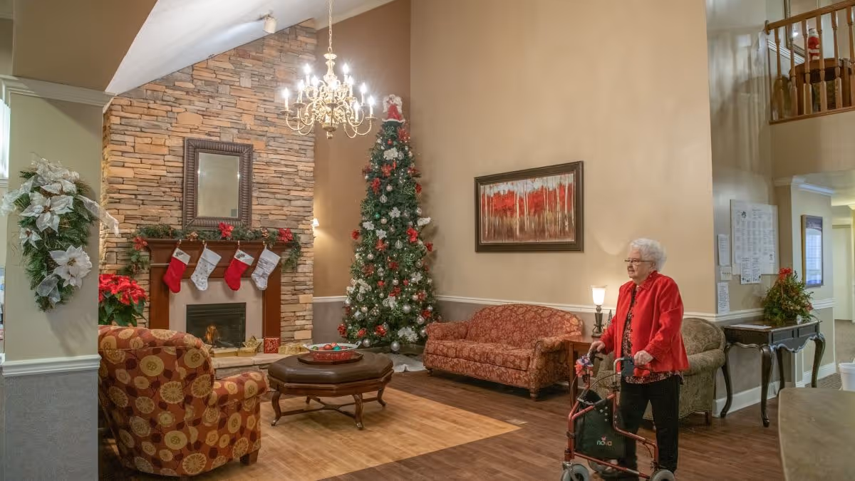 Interior common living area with a decorated Christmas tree, fireplace, seating, chandelier, and an elderly woman using a walker.